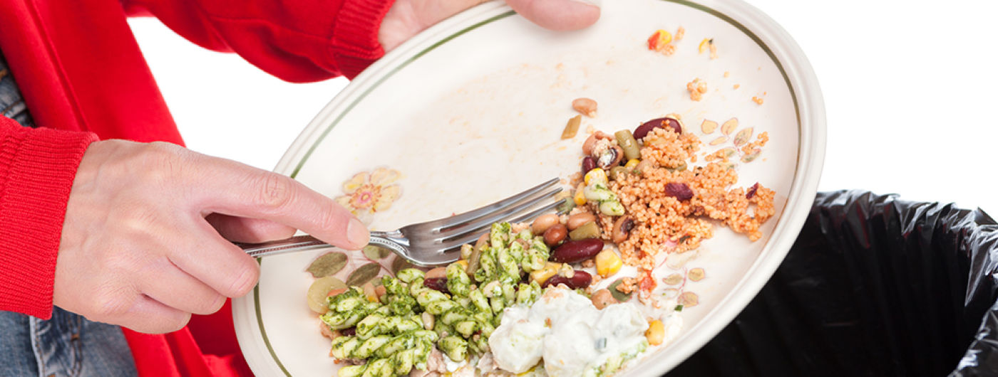 Close-up of a woman sweeping the leftovers from a meal into a domestic garbage bin. The background is pure white.